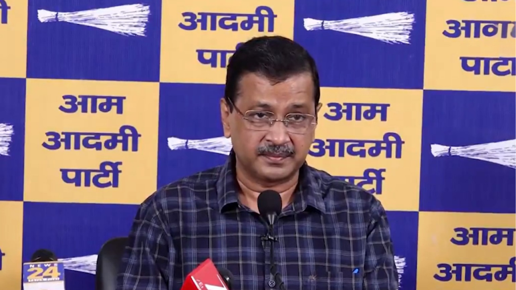 Man with glasses speaks into a microphone at a press conference, seated before a blue-yellow Aam Aadmi Party backdrop.