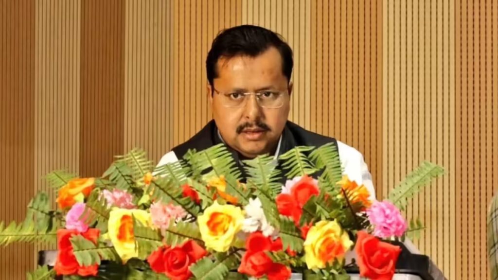 Man wearing glasses and a black vest speaks at a podium adorned with colorful flowers in front of a wooden panel backdrop