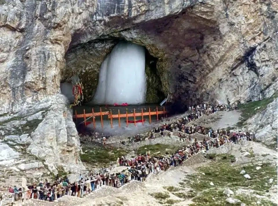 Long queue of people waiting outside a cave shrine with a large white frozen fountain inside the cave.