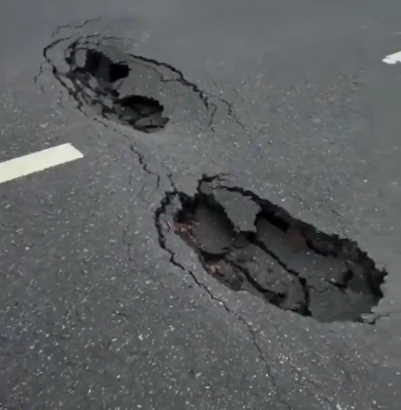 Screenshot 2026-07-02 132529 - The Journalist News Two large potholes in a cracked asphalt road with a white lane stripe nearby, indicating severe road damage.