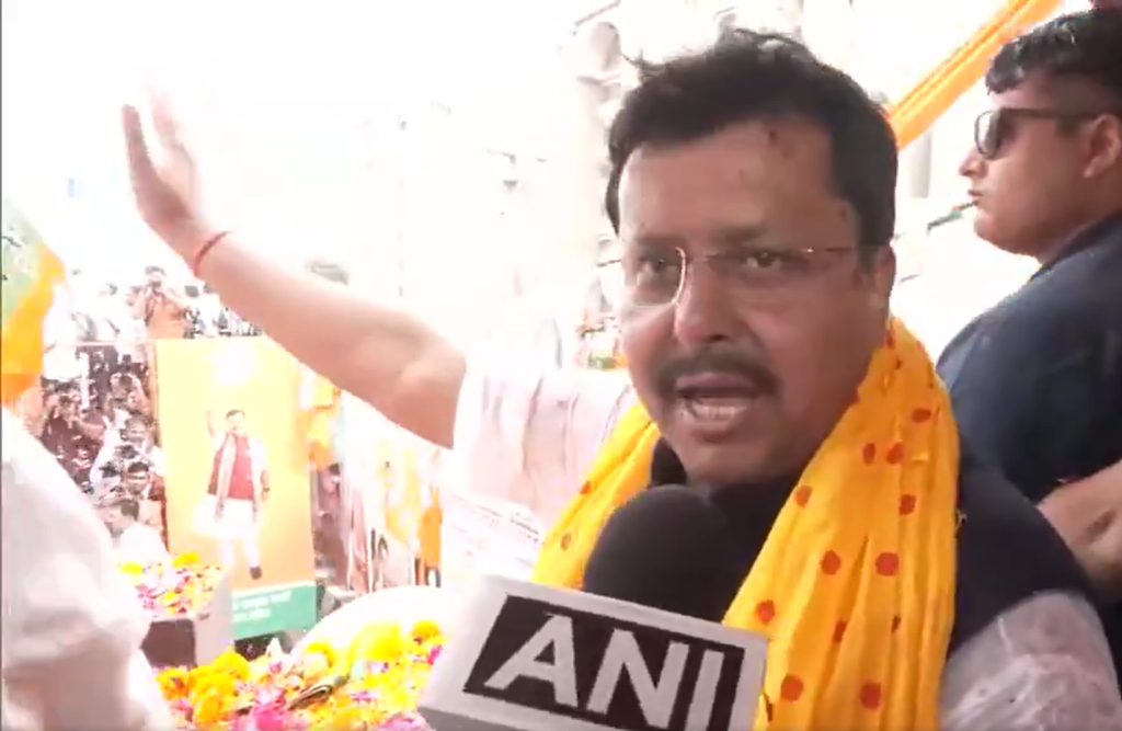 Man wearing glasses and a yellow scarf speaks into a microphone labeled ANI, gesturing with his right hand in a press interview.