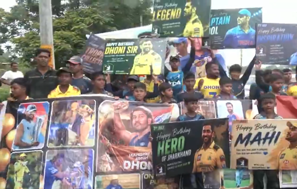 Screenshot 2026-07-07 155906 - The Journalist News Crowd of young fans holding up posters and banners celebrating a Dhoni birthday event, with images of Indian cricket stars in the background and signs reading 'Happy Birthday' and 'Hero of Jharkhand'.