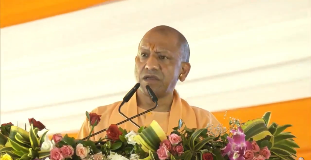 A Buddhist monk in an orange robe speaking at a podium adorned with pink and white flowers, with a microphone in front of him.