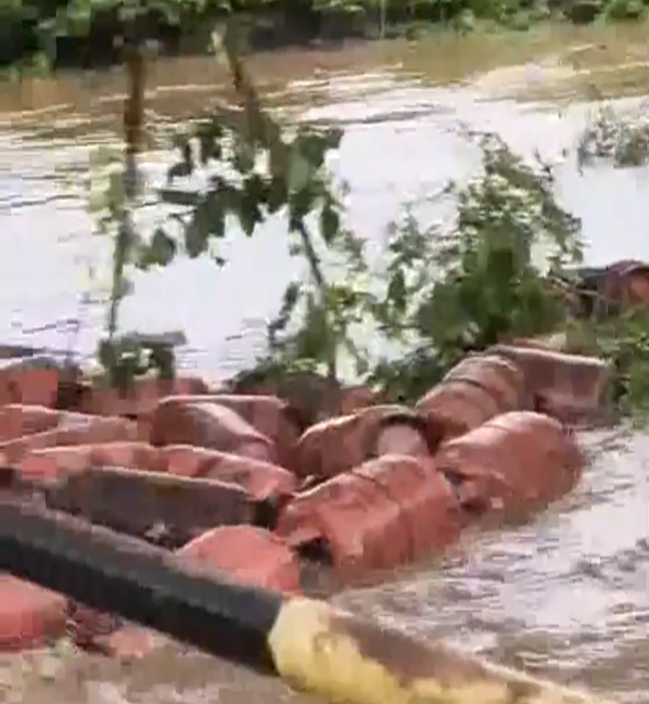 Screenshot 2026-07-09 150612 - The Journalist News Hose spraying water over a pile of red bricks or blocks along a flooded riverbank, with trees and water in the background (outdoor scene).