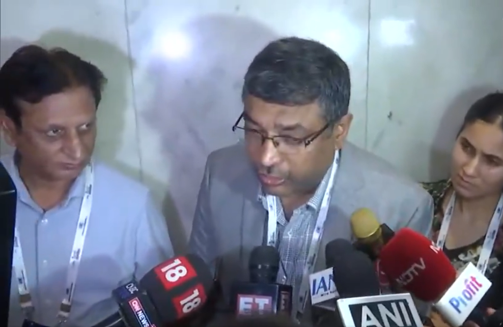Screenshot 2026-07-09 162103 - The Journalist News Man wearing a gray blazer and glasses speaks to reporters as multiple microphones surround him.