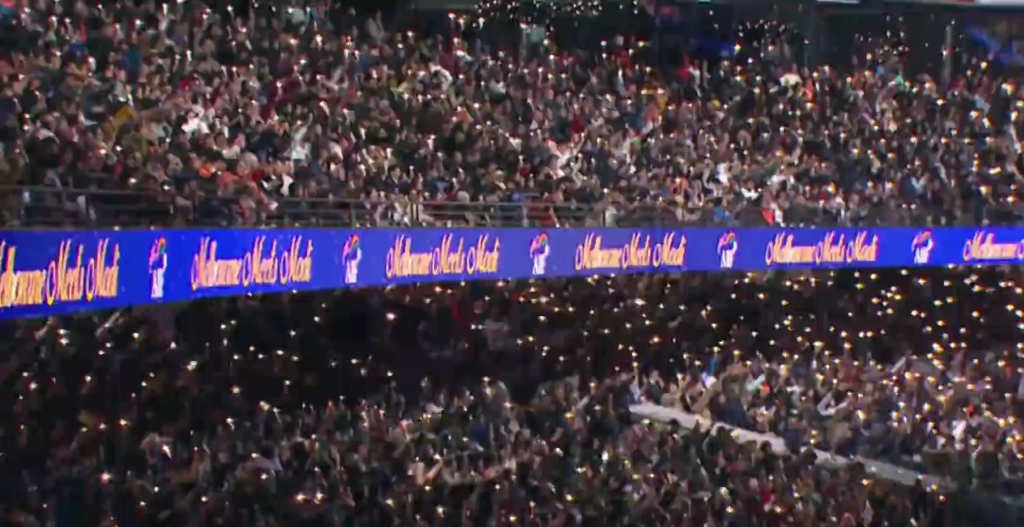 Large crowd of spectators in a packed stadium, many holding up phones with lights, behind a blue sponsor banner along the railing.