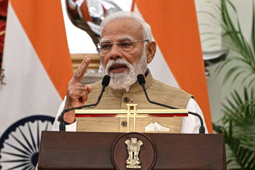 Speaker at a podium with two microphones, delivering a formal address with a national flag backdrop and emblem on the lectern.