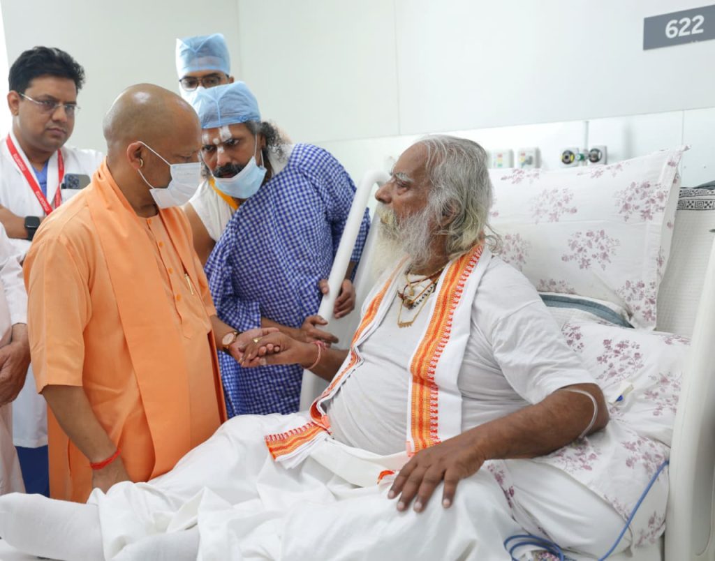 Senior man in orange robes greets an elderly patient in a hospital bed, shaking his hand as medical staff look on