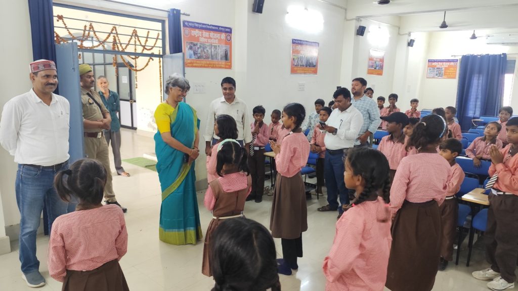 Teacher in a teal sari speaks to students in pink uniforms as adults observe in a bright classroom nearby.