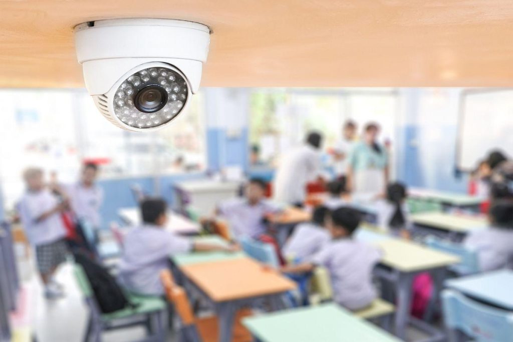 Security camera mounted over a colorful classroom filled with blurred students at desks in the background.