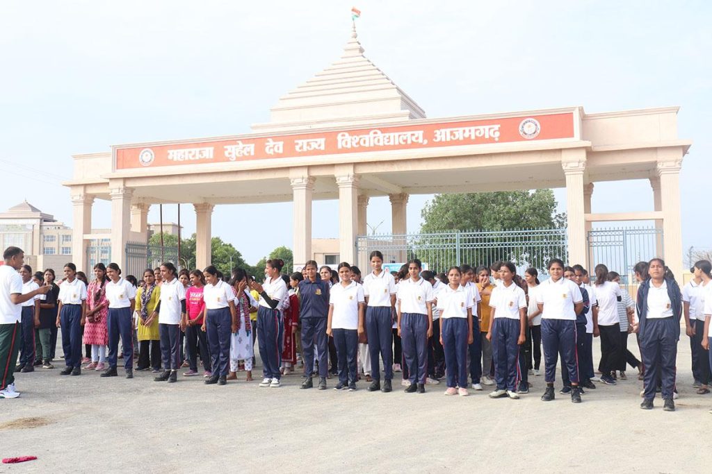 Group of students in white shirts and blue pants standing in a line outside a campus entrance with a red signboard in Hindi above the gate.