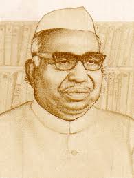 Portrait of a man wearing a white Gandhi cap and glasses, posed in front of a bookshelf.