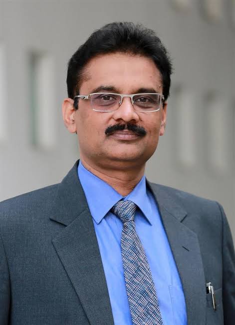 Portrait of a man in a gray suit, blue shirt, and patterned tie, wearing glasses against a neutral background.